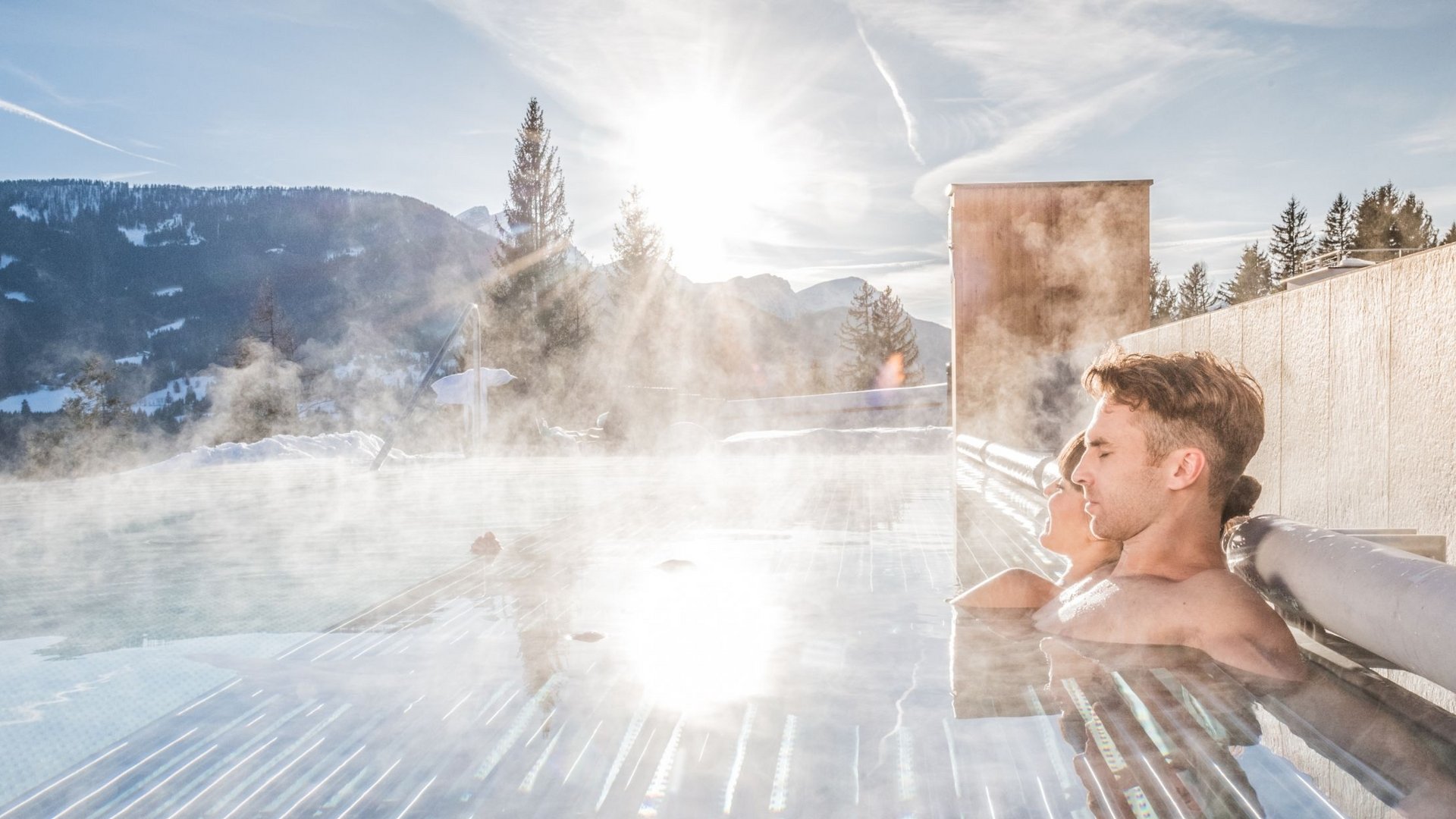 Paar entspannt im dampfenden Außenpool mit Bergblick bei Sonnenschein