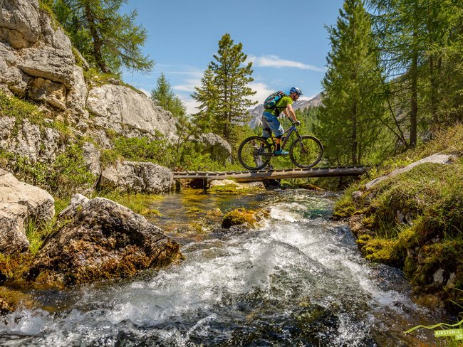 Alpen Tesitin: Ihr Bikehotel im Pustertal Mountainbiker überquert Holzbrücke über einen Bach in bewaldeter Berglandschaft
