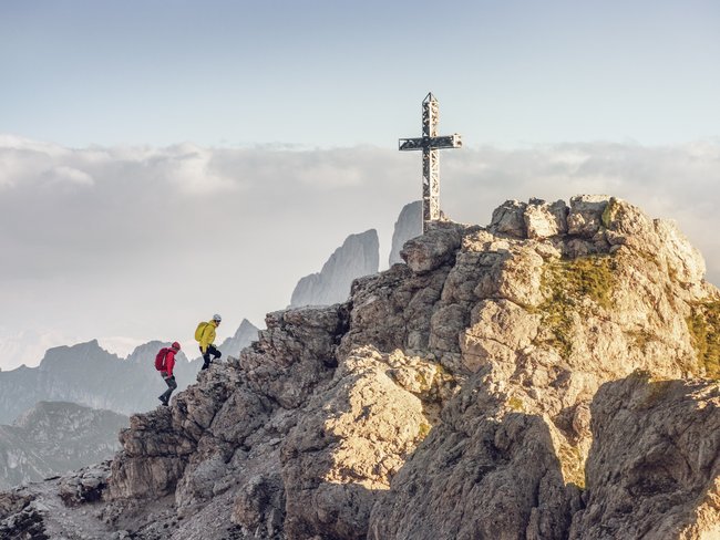 Zwei Wanderer erklimmen felsigen Gipfel mit großem Gipfelkreuz im Bergpanorama