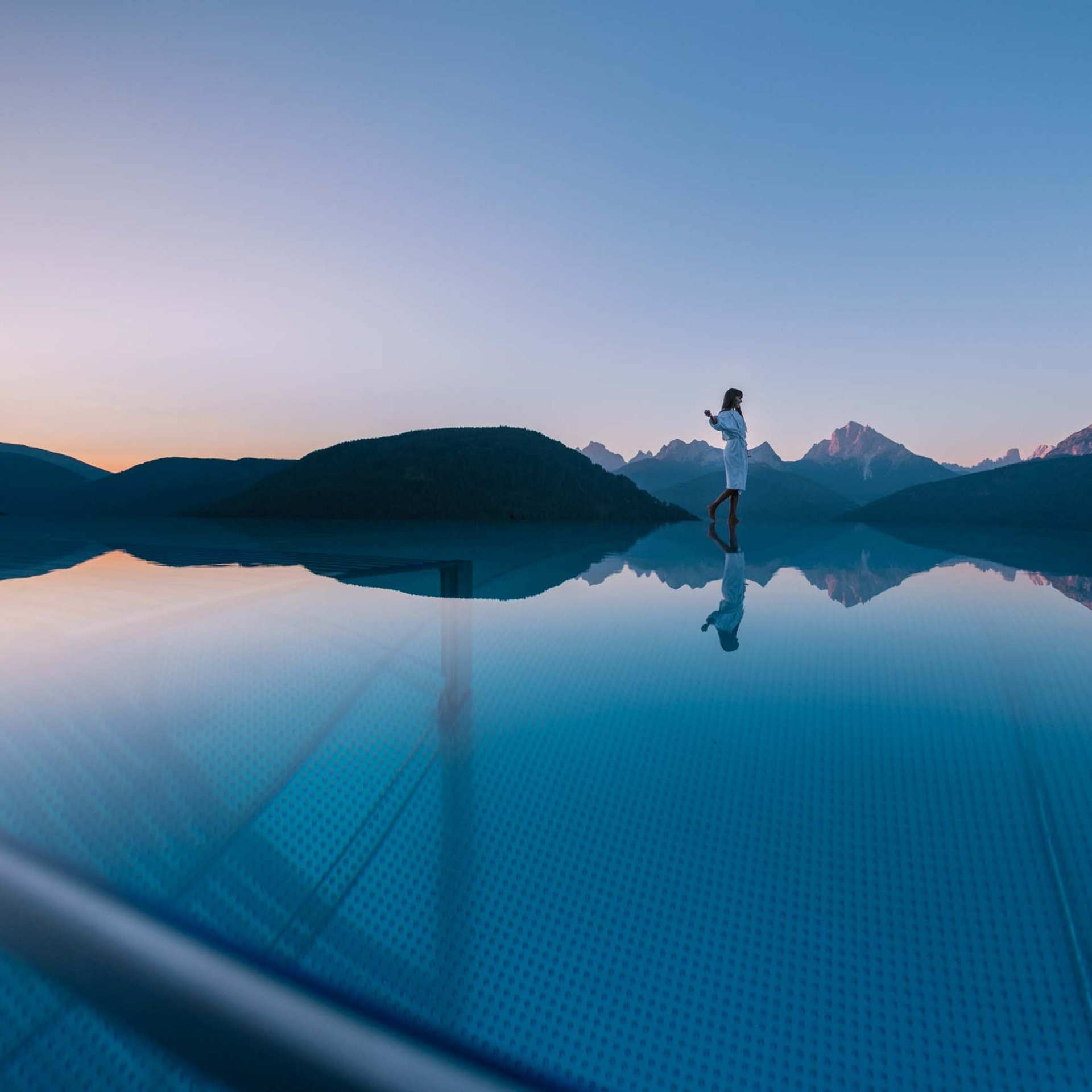AT Best of Frau steht am Rand eines Infinity-Pools mit Bergblick bei Sonnenuntergang