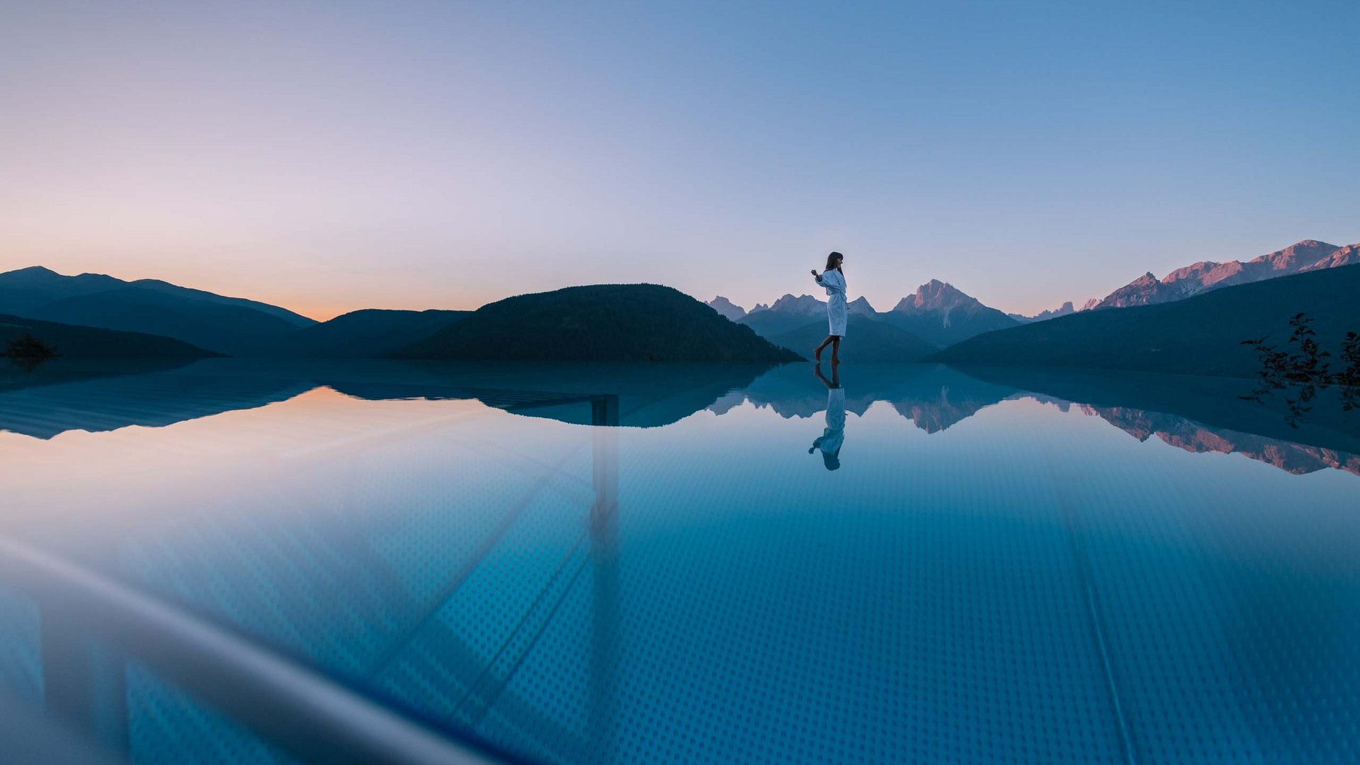 Frau steht am Rand eines Infinity-Pools mit Bergblick bei Sonnenuntergang
