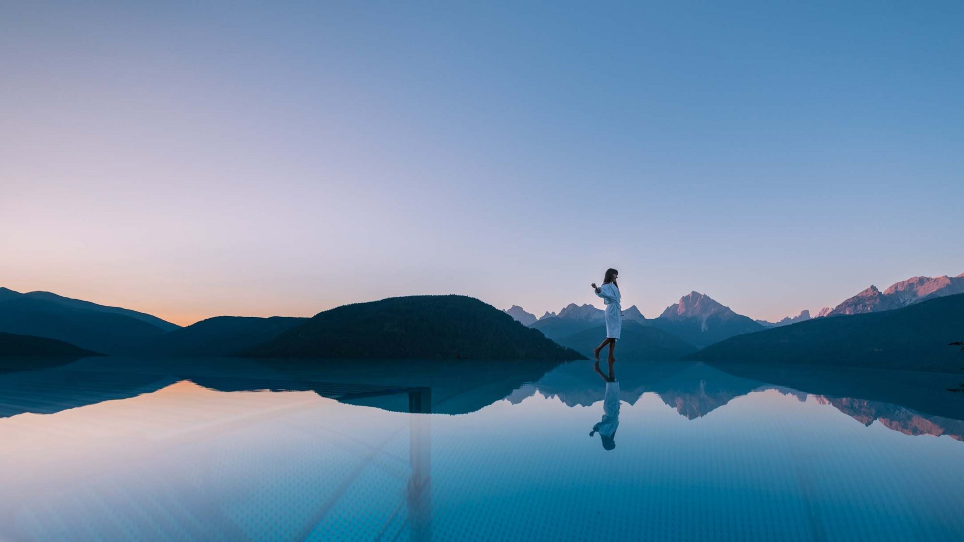 Angebote aus den Dolomiten Frau steht am Rand eines Infinity-Pools mit Bergblick bei Sonnenuntergang