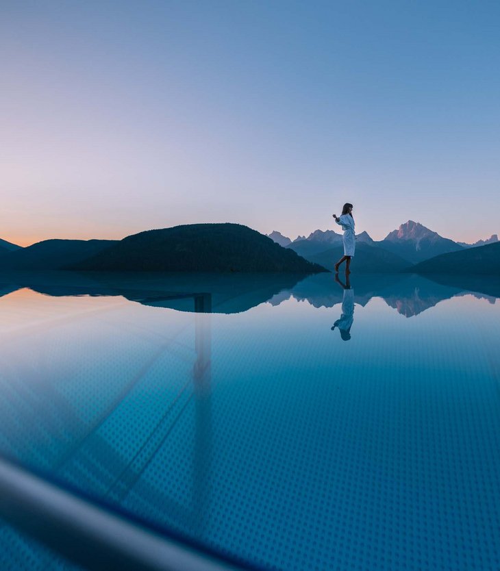 Your 5-star hotel in Val Pusteria/Pustertal: Alpen Tesitin Woman standing at edge of infinity pool with mountain view at sunset