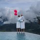 Paar in Bademänteln mit zwei roten Luftballons an einem Pool vor nebligen Bergen