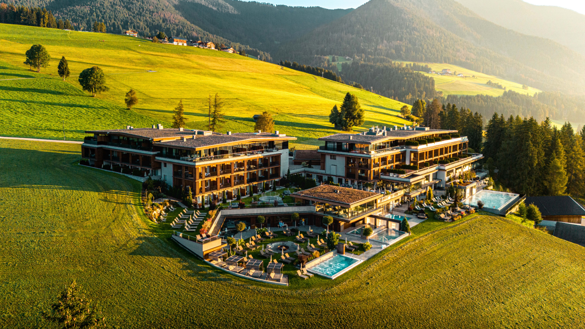 Luxury hotel with pools on green hill against forested mountains at sunset