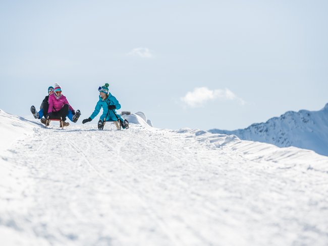 Vacanze invernali in Val Pusteria all’Alpen Tesitin Tre persone che slittano su una pista innevata sotto il sole