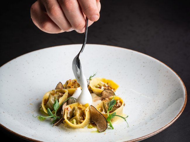 Hand mit Löffel gibt weiße Sauce auf gefüllte Pasta mit Trüffeln