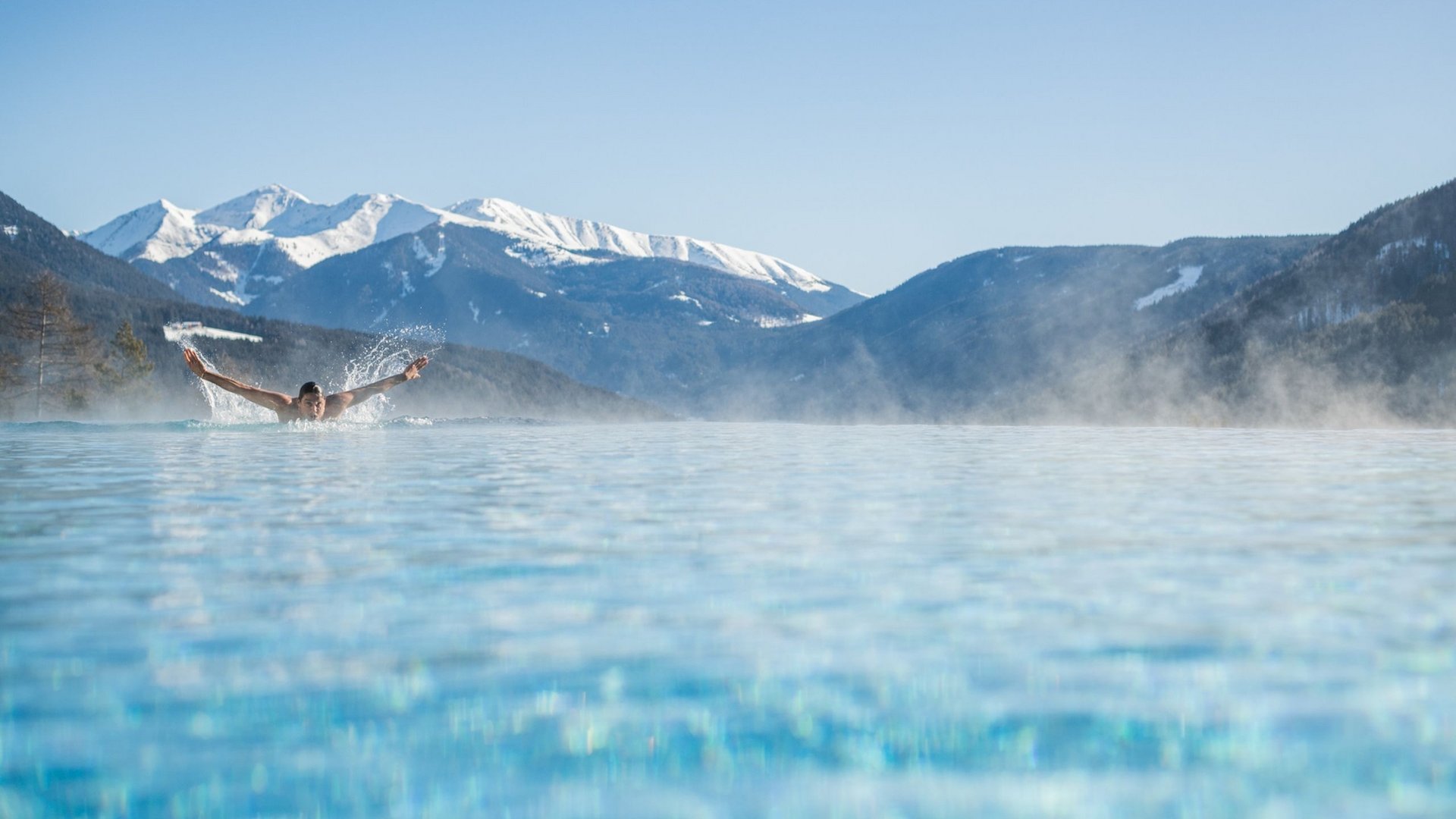 Person schwimmt in beheiztem Pool vor verschneiten Bergen