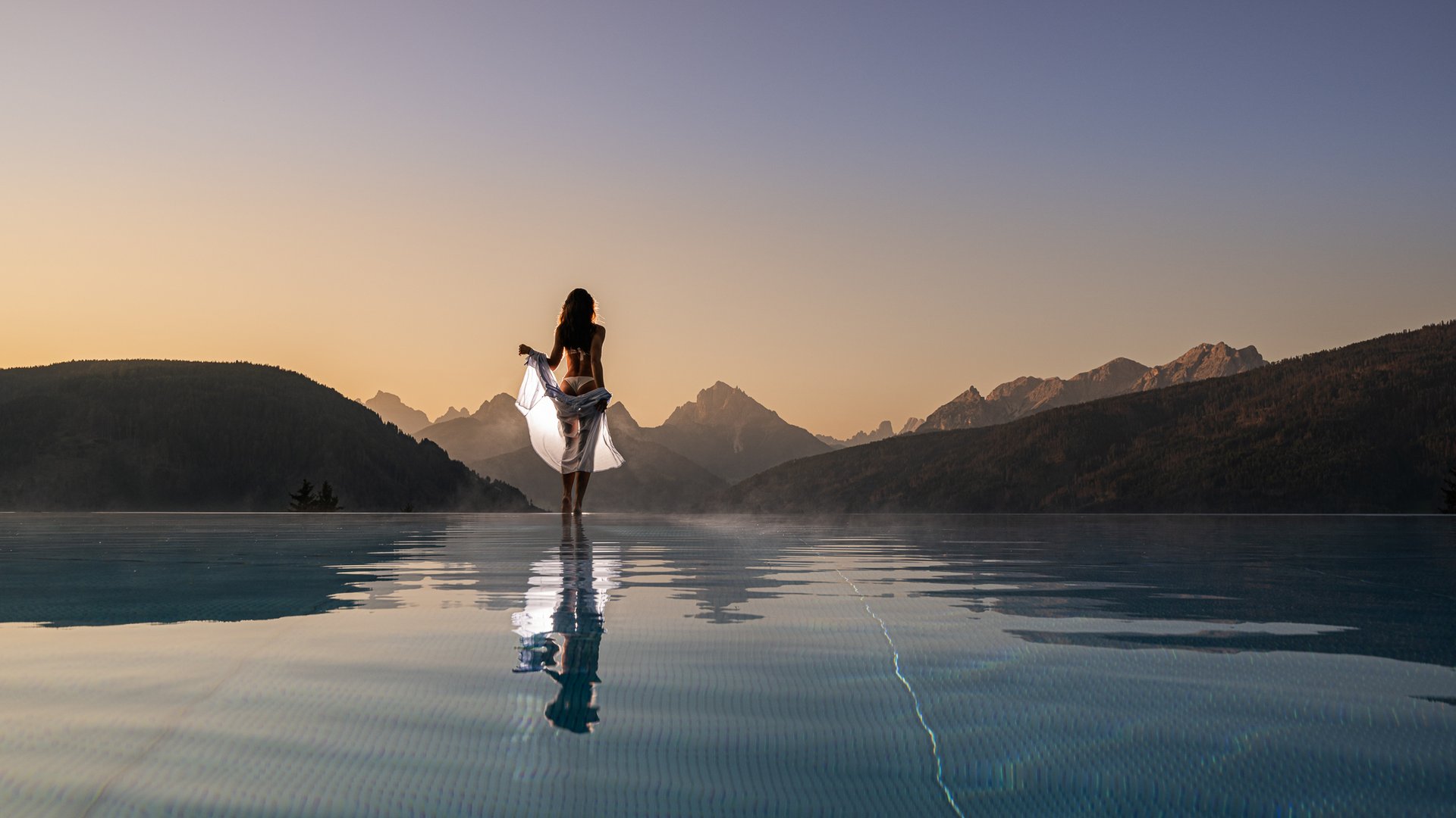 Woman in white cloth at infinity pool with mountain view at sunset