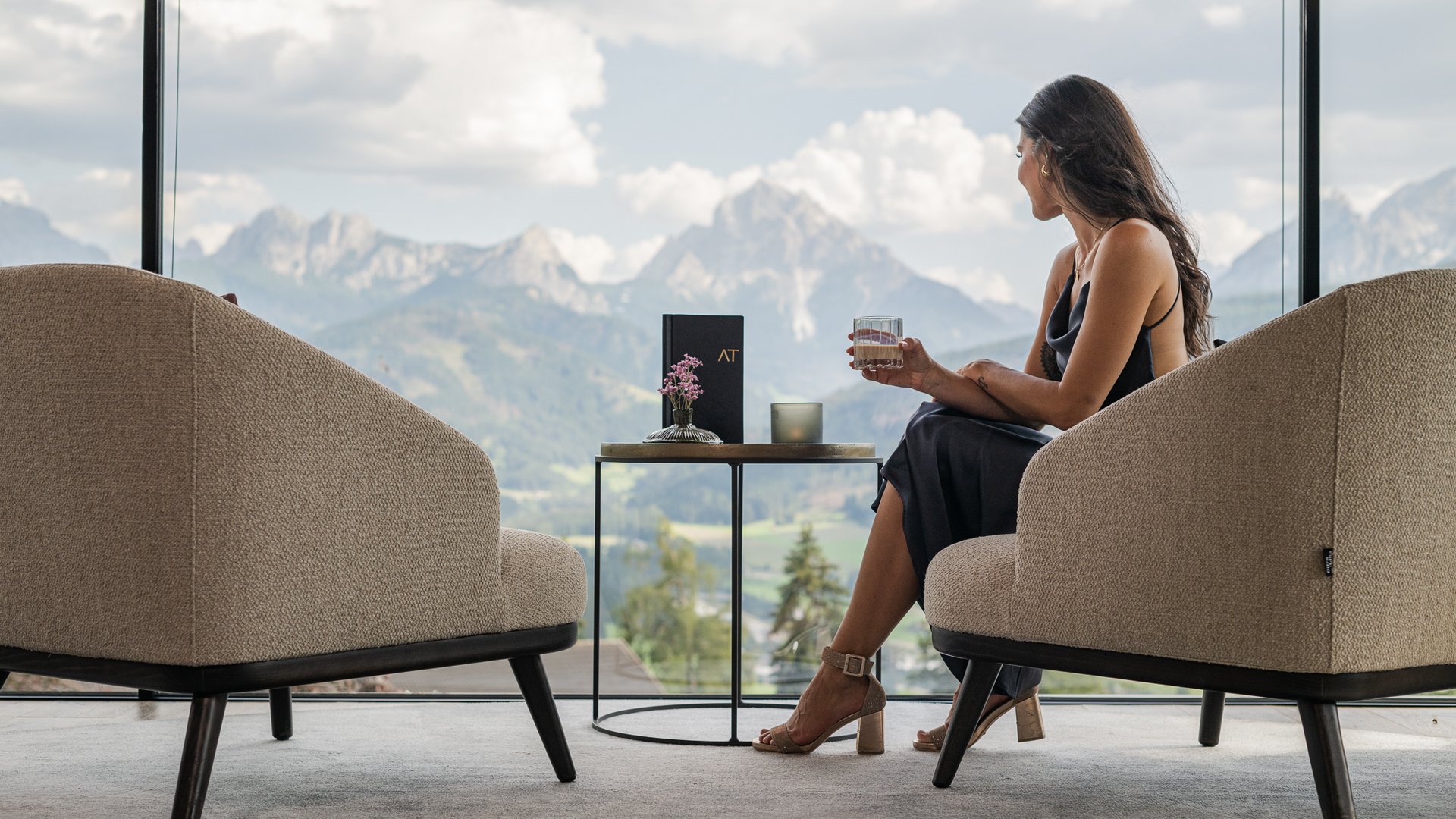 Woman in elegant dress sitting indoors with mountain view holding half-full glass