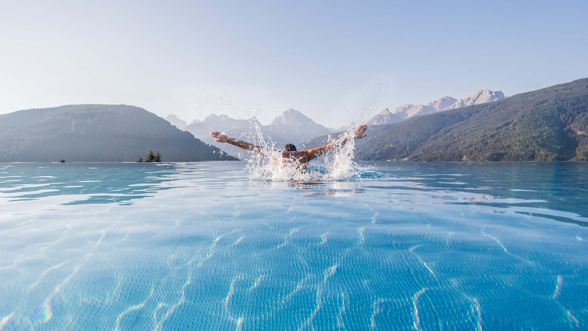 Your hotel in Val Pusteria/Pustertal with pool oasis Man swimming in infinity pool with clear mountain view on sunny day