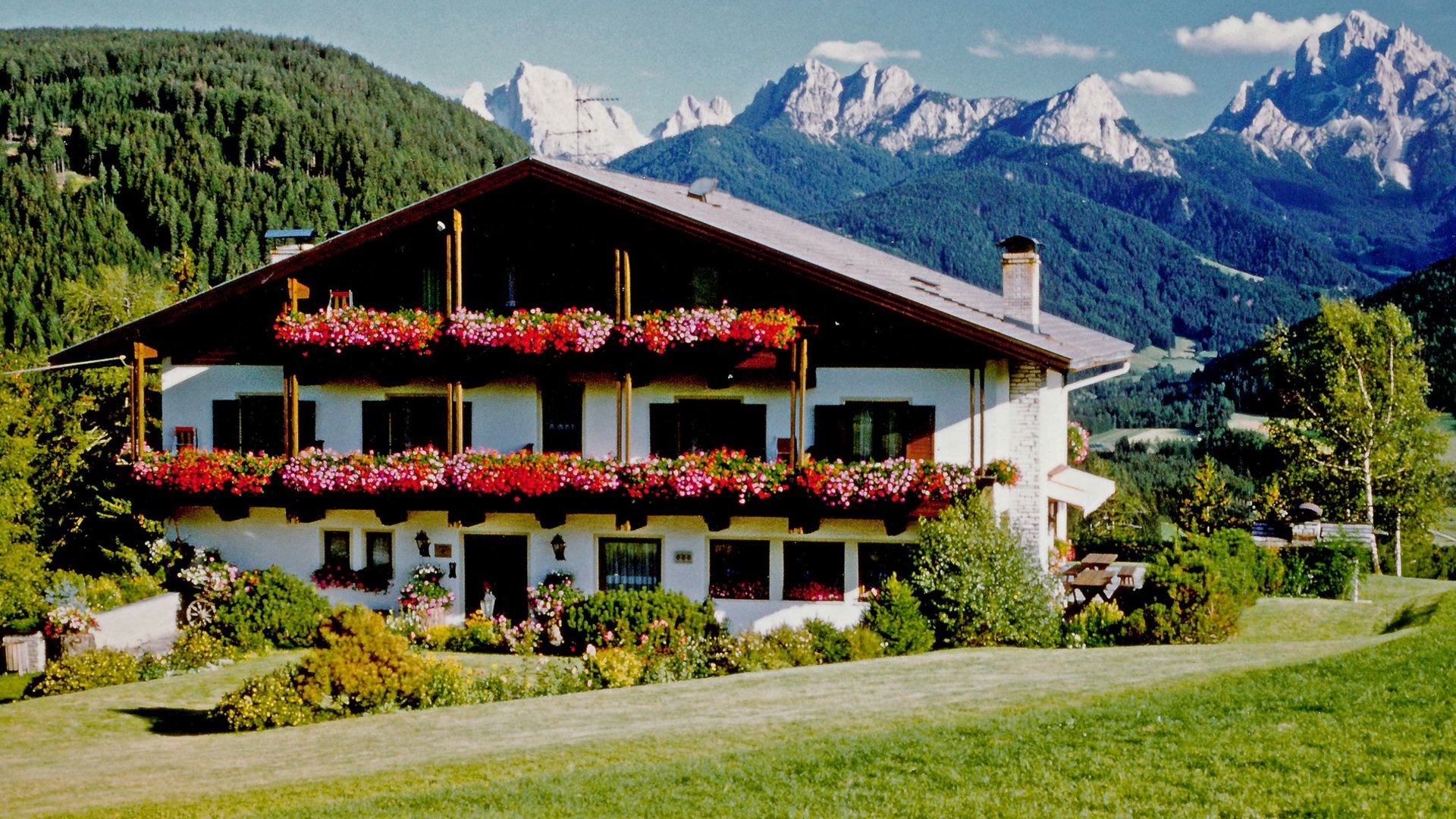 Casa alpina con balconi fioriti davanti a montagne boscose e cielo azzurro