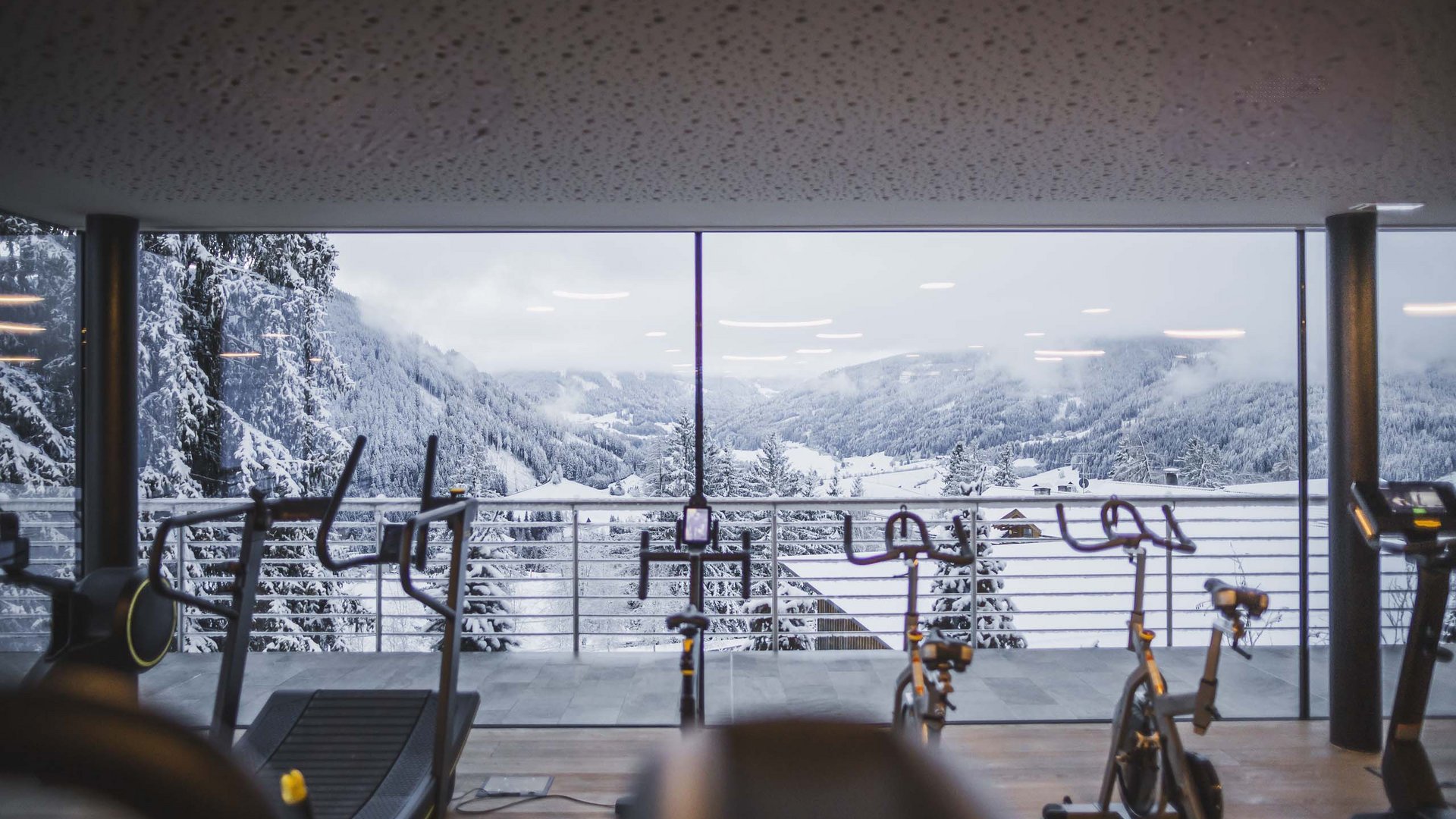 Fitnessraum mit Fahrrädern vor Panoramafenster und verschneiter Berglandschaft