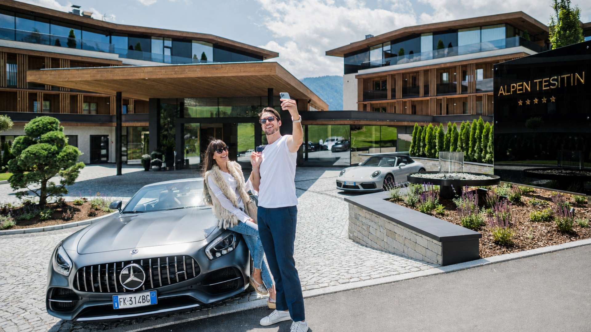 Rent a sports car in South Tyrol Couple taking a selfie outside a modern hotel with luxury cars and Alpen Tesitin sign