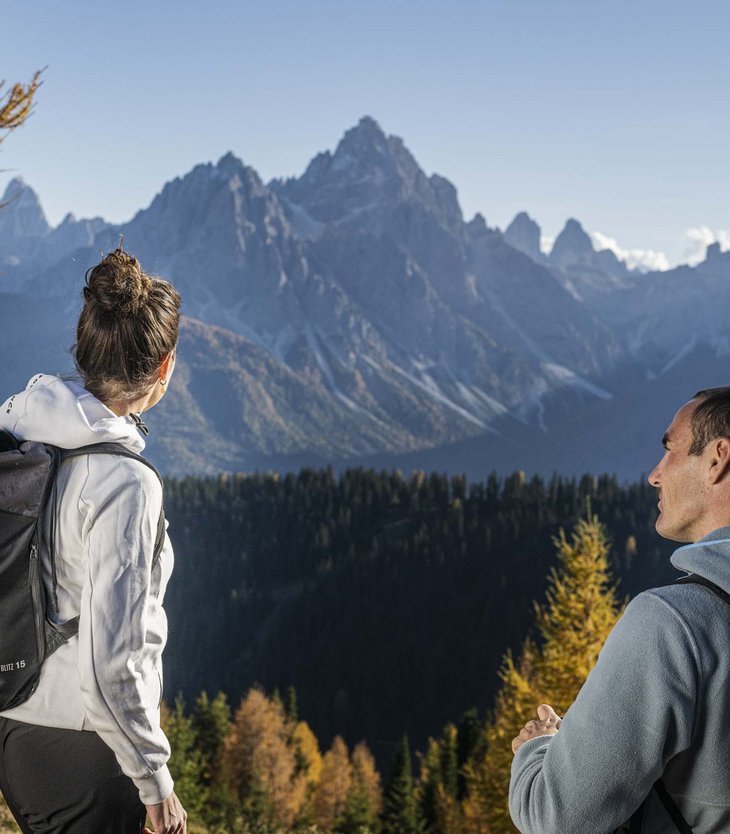 Due escursionisti con zaini guardano le cime montuose e la foresta autunnale.