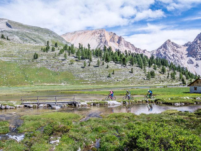 Drei Radfahrer auf einem Bergpfad mit Alpenlandschaft und Hütte