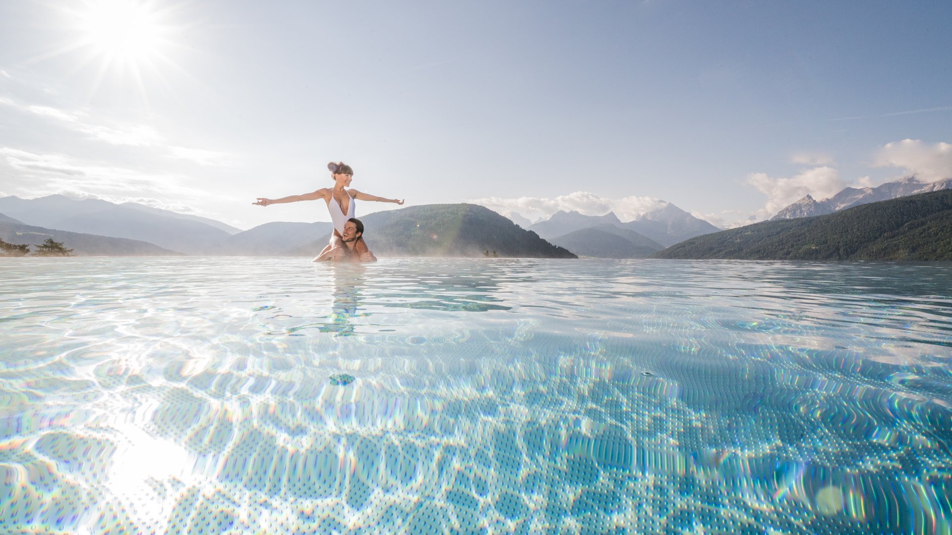 Your hotel in Val Pusteria/Pustertal with pool oasis Couple in infinity pool with mountain view on a sunny day