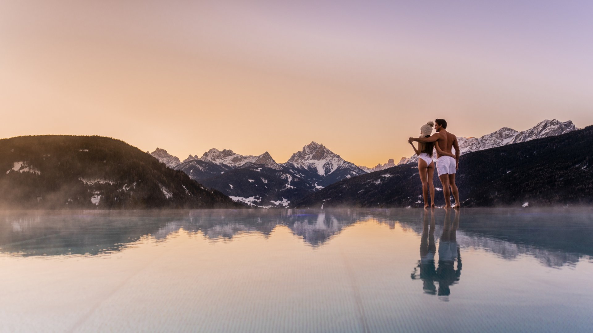 Il vostro hotel in Val Pusteria con piscina: la vostra oasi Coppia in costume da bagno ai bordi di una piscina con montagne al tramonto