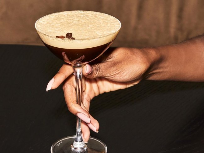 Hand hält ein Cocktailglas mit einem Espresso Martini auf schwarzem Tisch