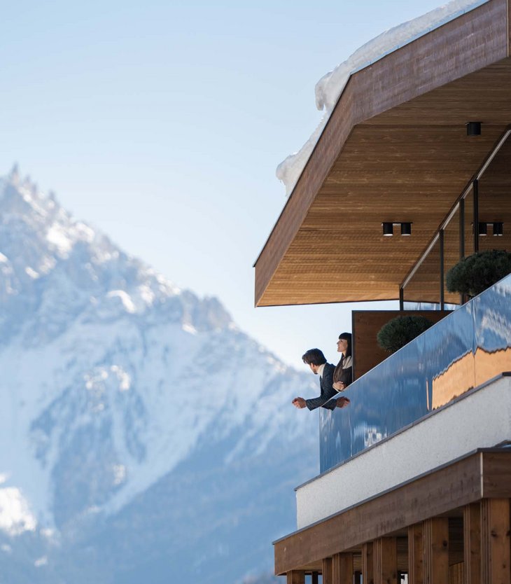 Your 5-star hotel in Val Pusteria/Pustertal: Alpen Tesitin Couple on balcony of modern chalet looking at snow-covered mountain