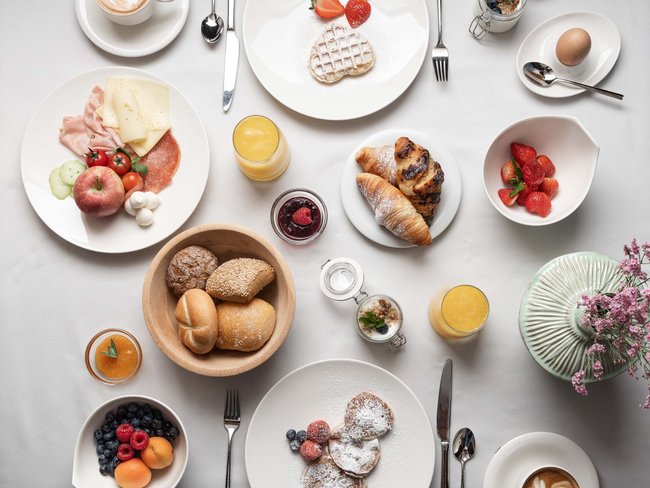Verschiedene Frühstücksgerichte auf weißem Tisch mit Kaffee, Obst und Gebäck