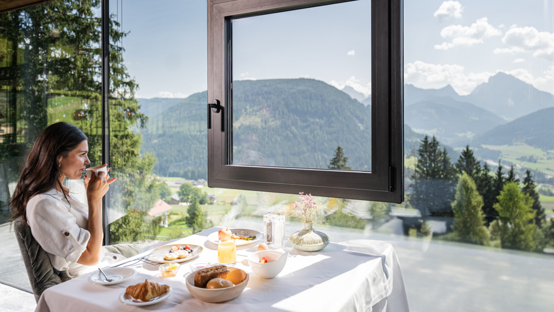 Frau trinkt Kaffee bei Frühstück mit Bergblick durch großes Fenster