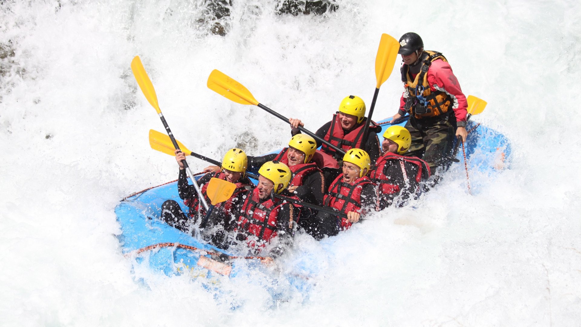 Gruppo che fa rafting in acque bianche con caschi gialli e giubbotti rossi su gommone blu