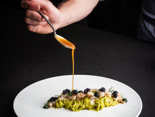 Ein Koch gießt Sauce auf kunstvoll angerichtete Pasta mit Meeresfrüchten und Kaviar.