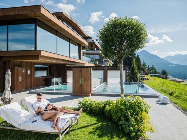 Paar entspannt sich auf Liegen vor modernem Holzhaus mit Bergblick und Pool