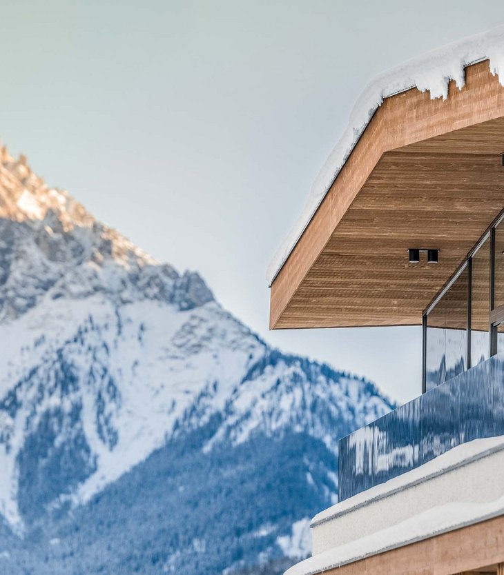Moderne Hütte mit Holzfassade und Glasgeländer vor verschneitem Berg