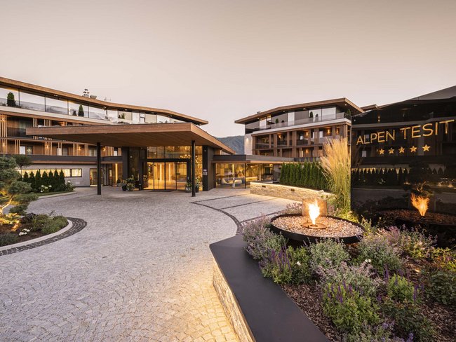 Urlaub im Hotel in den Dolomiten Eingang eines modernen Hotels mit Feuerstelle und Alpenhintergrund bei Abenddämmerung