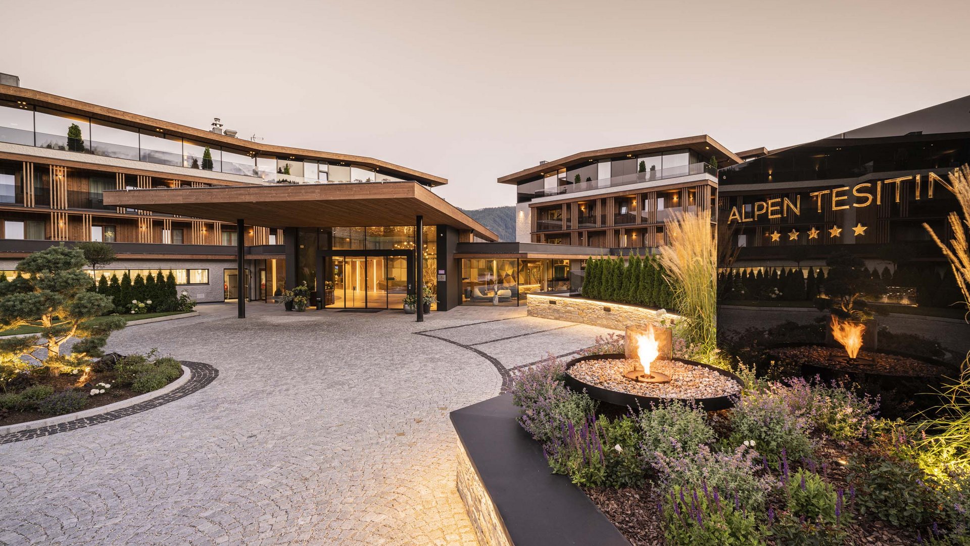 Entrance of a modern hotel with fire feature and alpine background at dusk