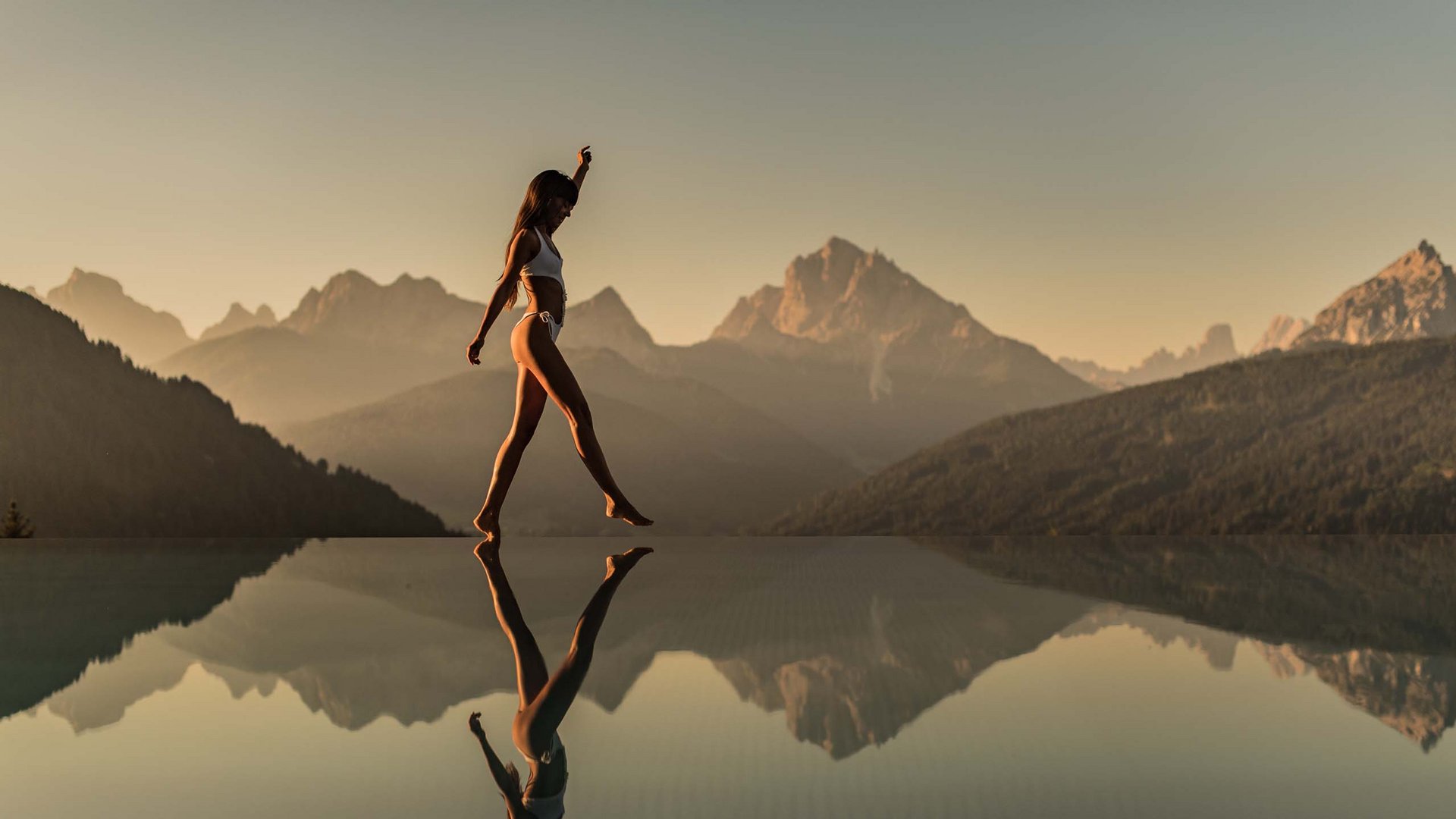 Frau in Badeanzug geht am Rand eines Pools mit Bergspiegelung bei Sonnenuntergang
