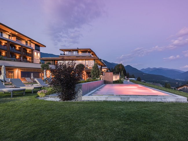 Complesso alberghiero moderno con piscina esterna illuminata e vista sulle montagne al crepuscolo