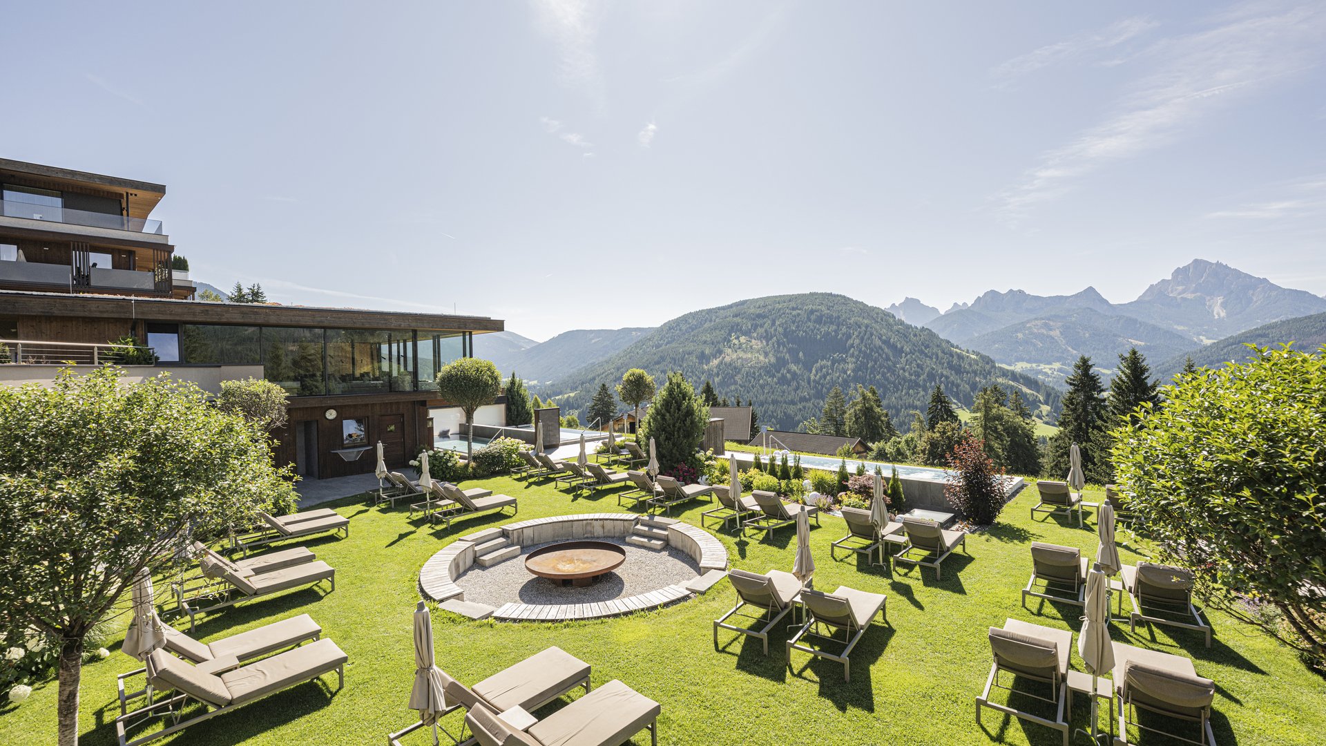 Hotel mit Sauna in Südtirol: Alpen Tesitin Sonnenliegen rund um ein Feuerbecken mit Bergkulisse und modernem Haus