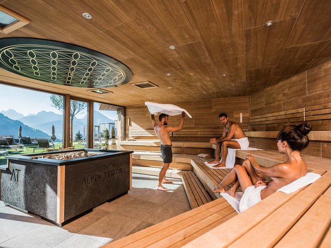 Tre persone rilassate in una sauna di legno con vista sulle montagne e grandi finestre