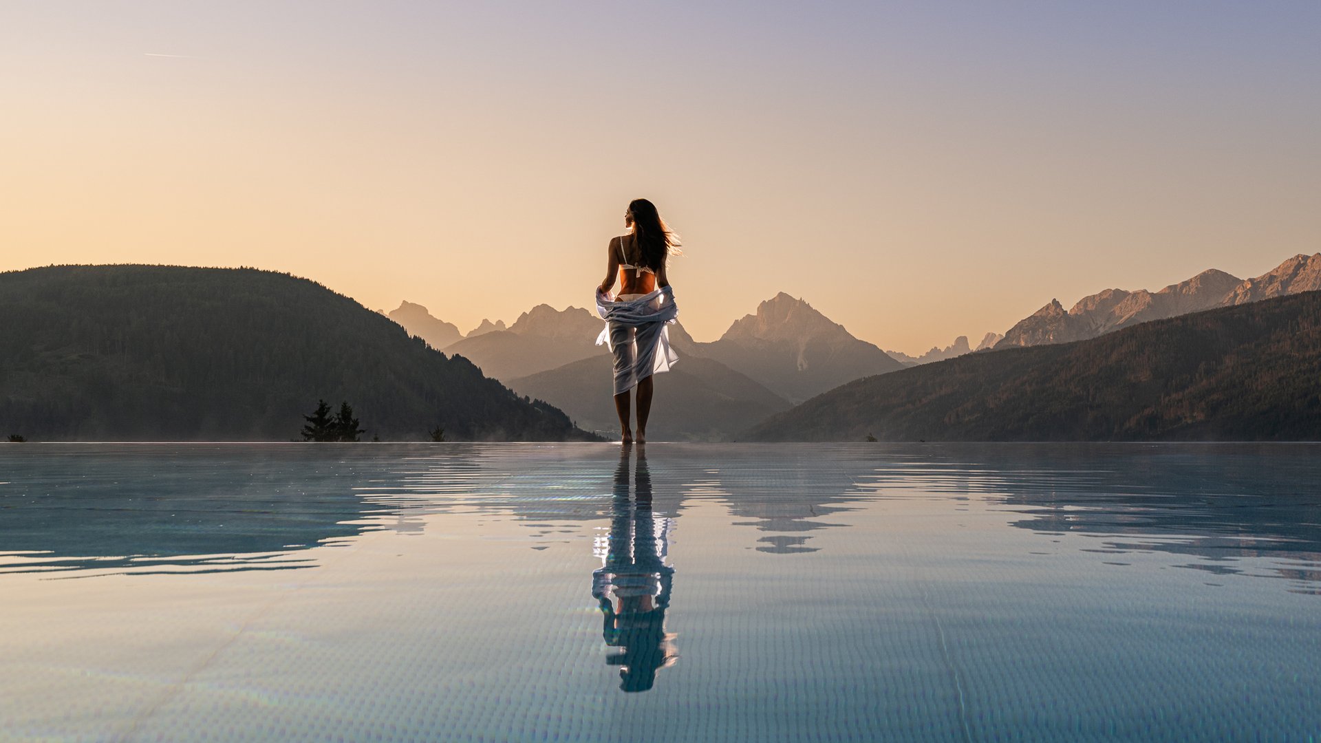 Your hotel in Val Pusteria/Pustertal with pool oasis Woman at infinity pool overlooking mountains at sunset
