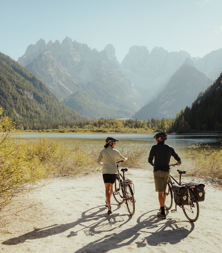 Zwei Radfahrer mit Helmen neben Bergsee und Bergen an sonnigem Tag
