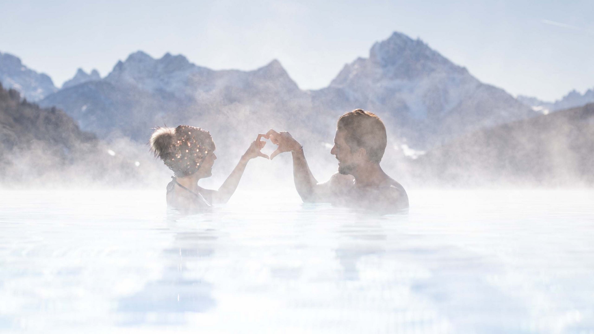 Angebote aus den Dolomiten Paar in heißem Pool vor Bergkulisse formt Herz mit Händen