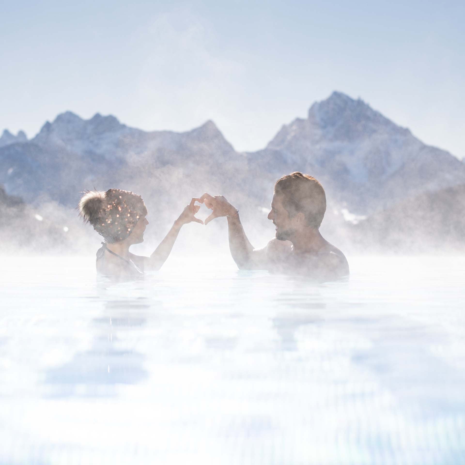 Your 5-star hotel in Val Pusteria/Pustertal: Alpen Tesitin Couple in hot pool with mountain backdrop making a heart with hands