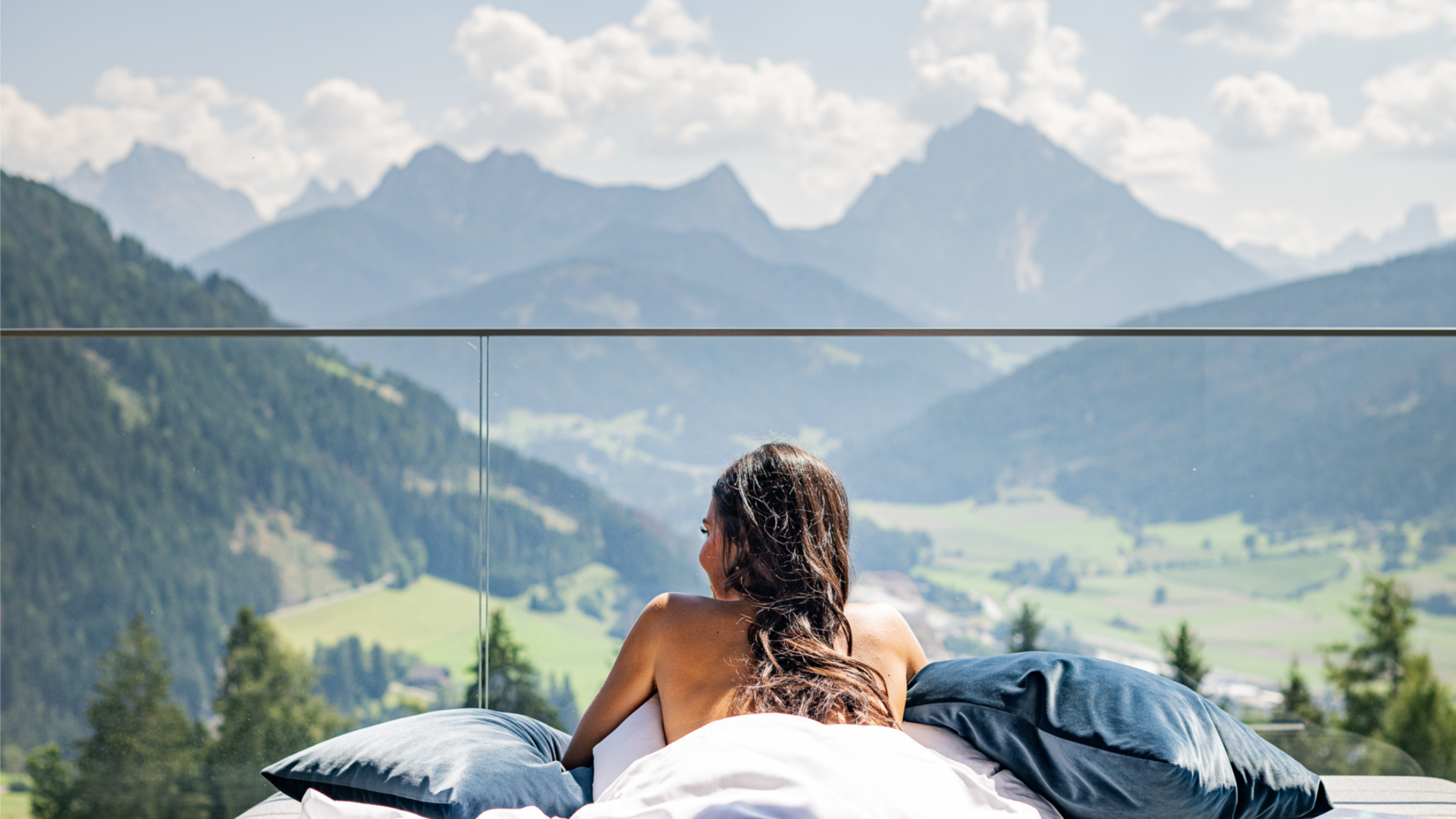 Spontanurlaub im Pustertal Frau liegt entspannt auf Bett mit Bergpanorama und Glasgeländer im Hintergrund