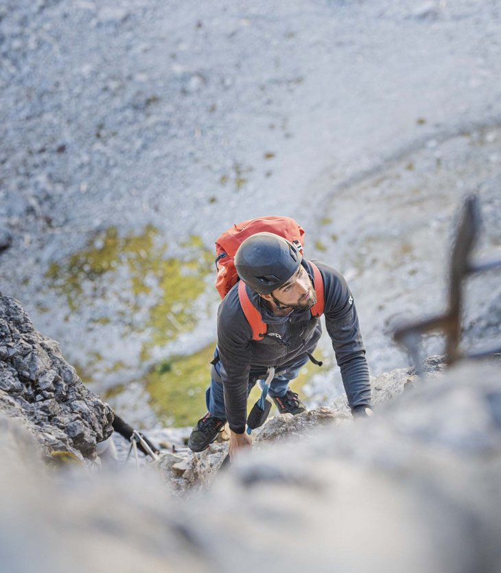 Mann mit Helm und Rucksack klettert an steiler Felswand hinauf
